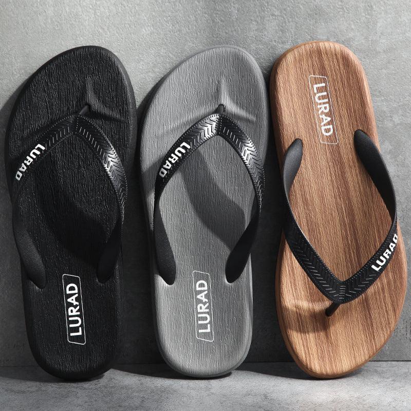 Three LURAD flip-flops in black, gray, and wood grain with textured soles and black straps on gray surface