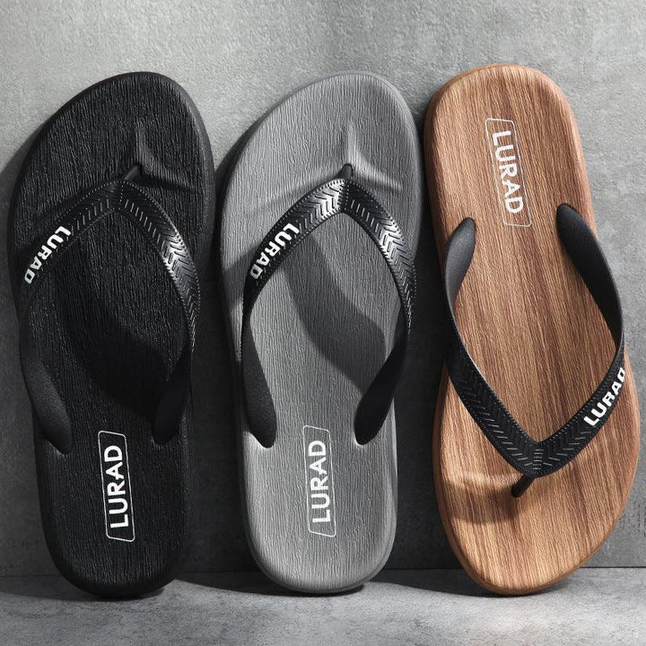 Three LURAD flip-flops in black, gray, and wood grain with textured soles and black straps on gray surface