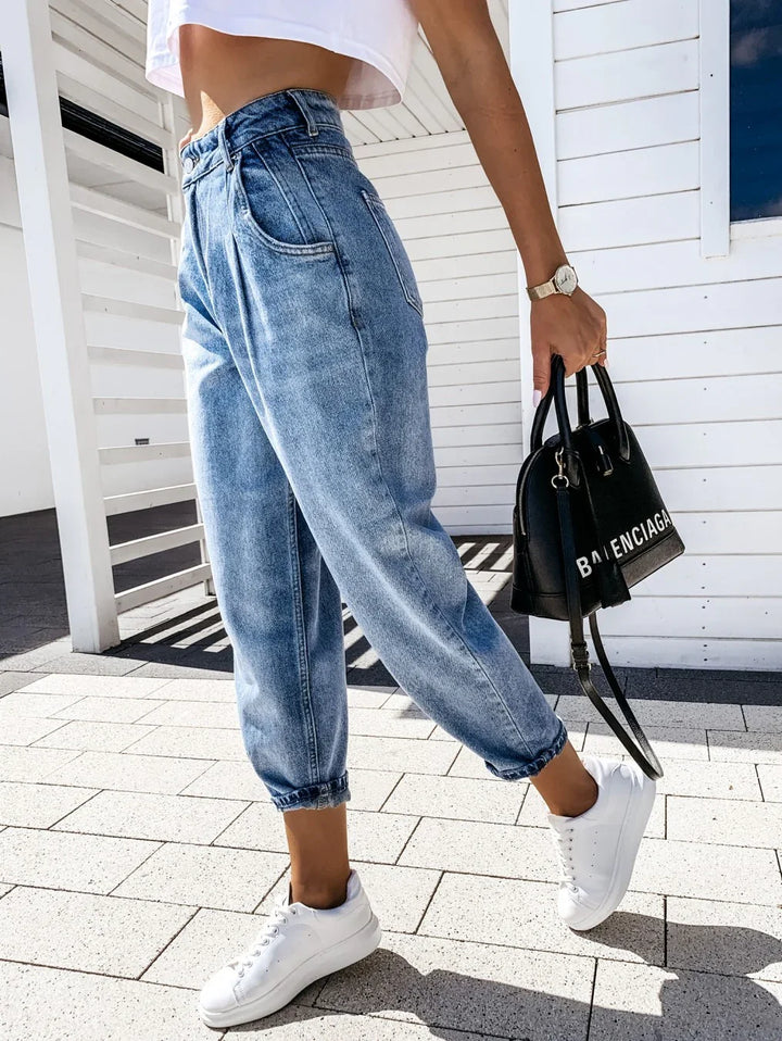 Woman wearing high-waisted light blue denim jogger jeans, white sneakers, white crop top, holding black Balenciaga handbag outdoors