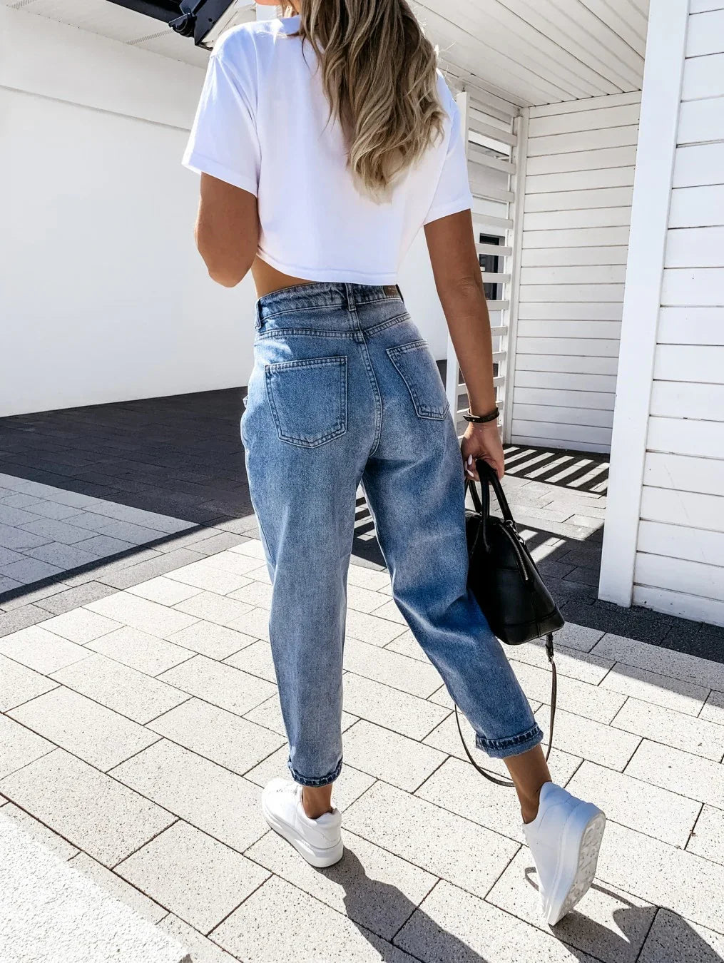 Woman walking outdoors wearing high-waisted blue jeans, white cropped t-shirt, white sneakers, and carrying black handbag