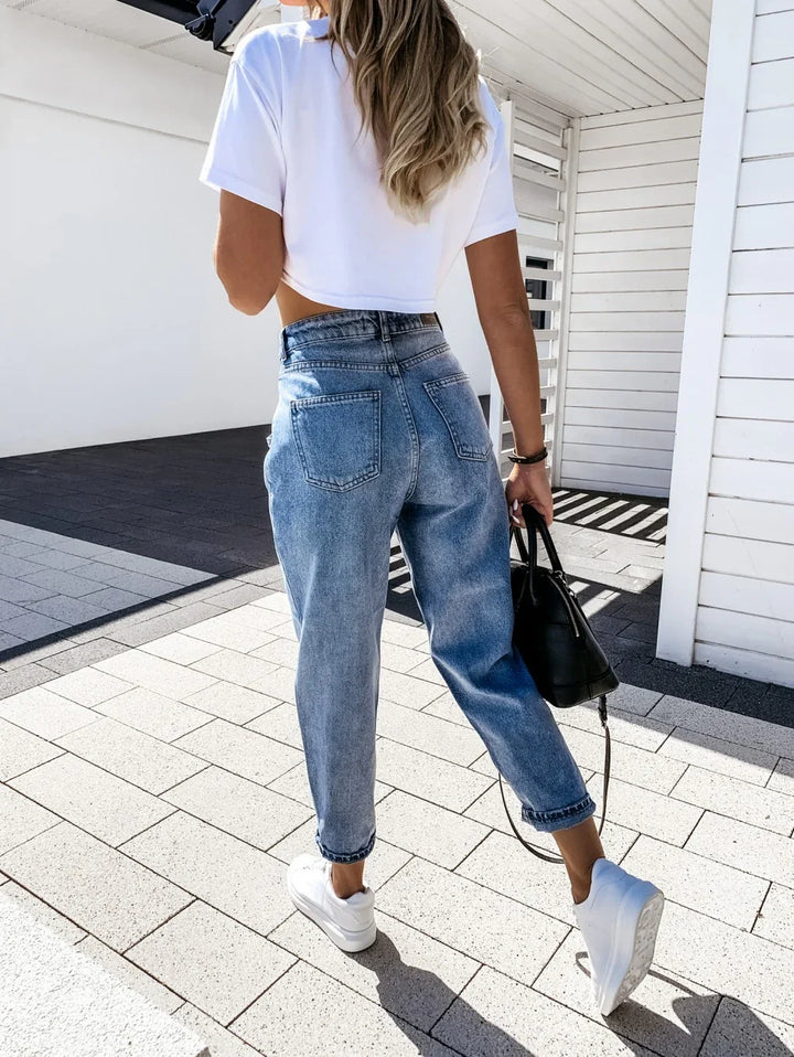 Woman walking outdoors wearing high-waisted blue jeans, white cropped t-shirt, white sneakers, and carrying black handbag