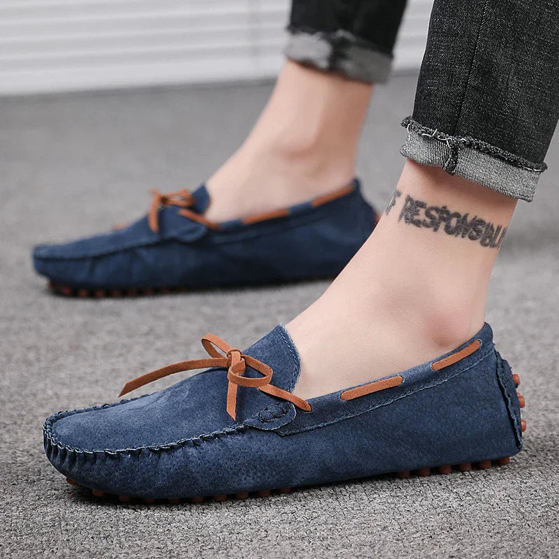 Feet wearing blue suede loafers with brown bow and accents, paired with rolled-up dark jeans