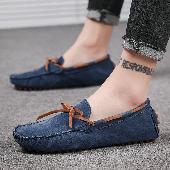 Feet wearing blue suede loafers with brown bow and accents, paired with rolled-up dark jeans