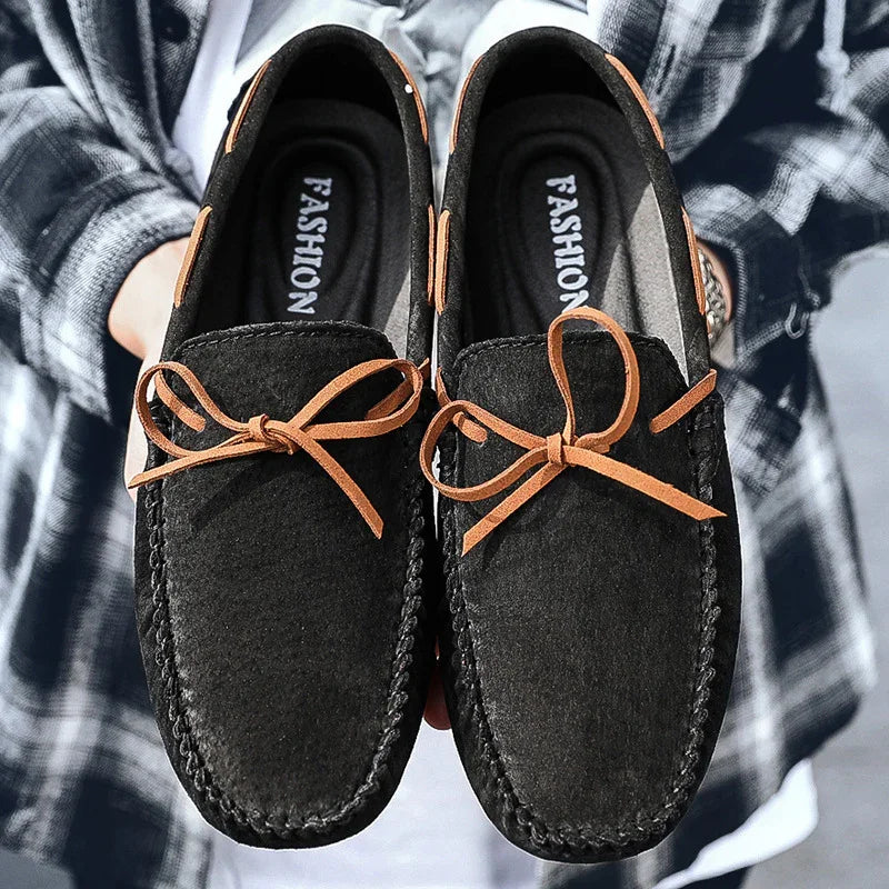 Pair of black suede loafers with brown leather bow laces held by person in plaid shirt
