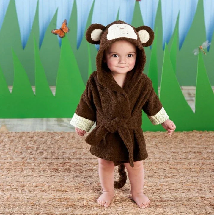 Toddler wearing a brown monkey costume bathrobe with hood and tail, standing on a textured mat with green grass backdrop