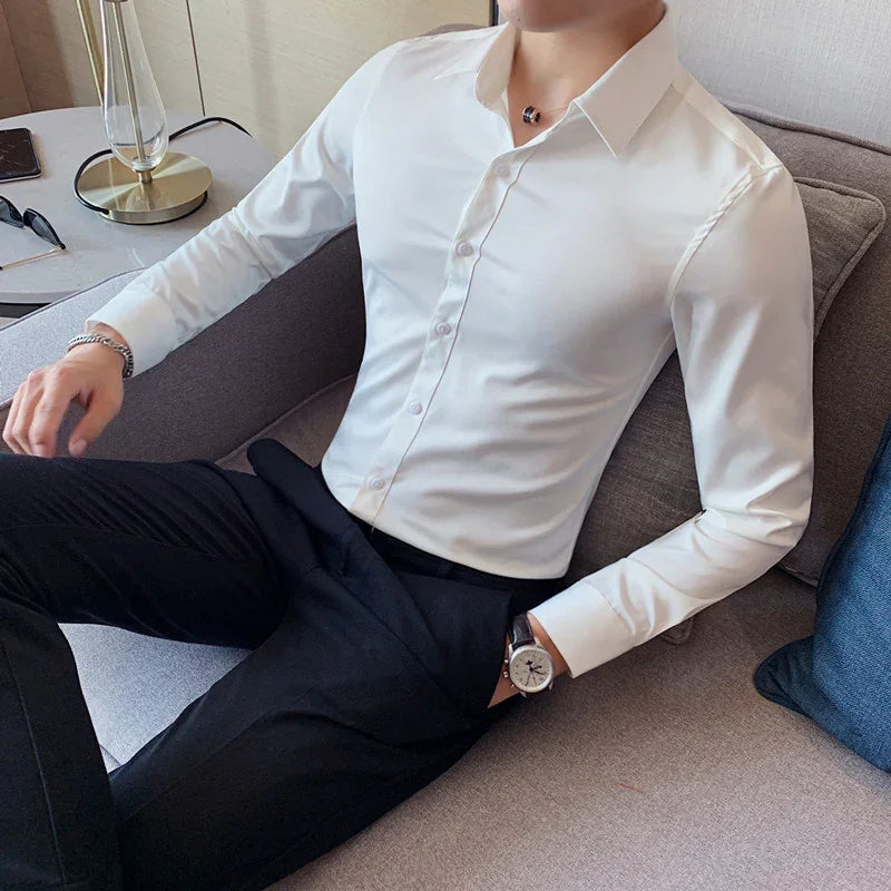 Man wearing a fitted white dress shirt and black pants sitting on a gray sofa with a watch on wrist