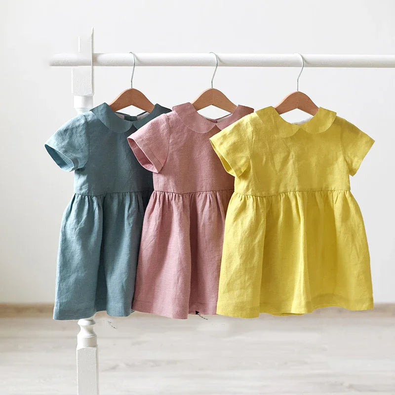 Three short-sleeve baby dresses in blue, pink, and yellow with Peter Pan collars hanging on wooden hangers