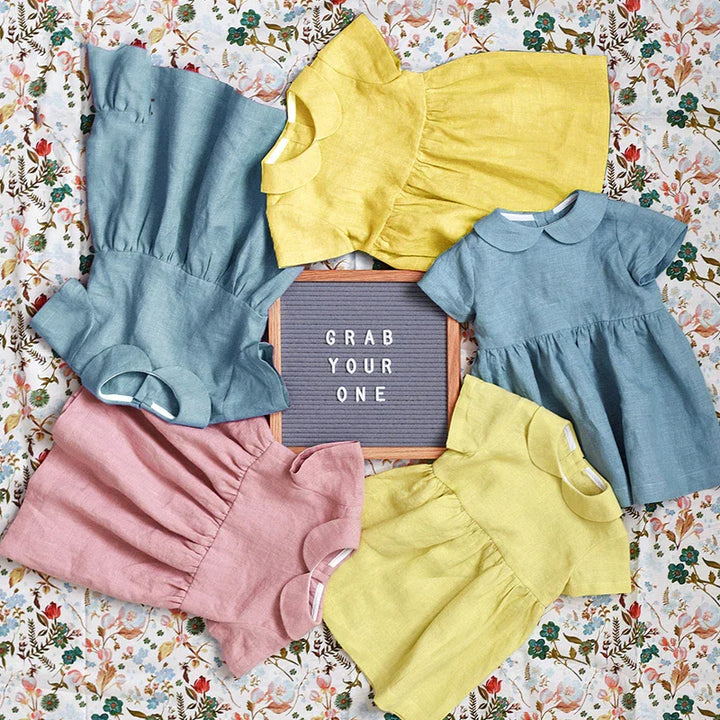 Flat lay of colorful toddler dresses in blue, yellow, and pink around a sign reading 'GRAB YOUR ONE' on floral background