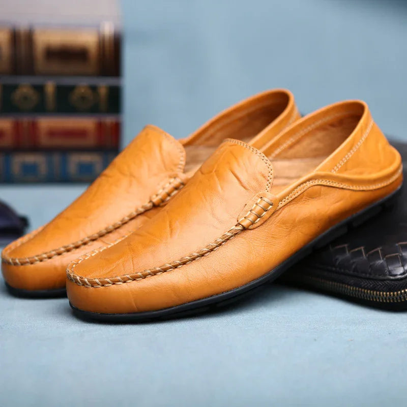 Pair of tan leather men's loafers with braided stitching on a light blue surface and books in the background