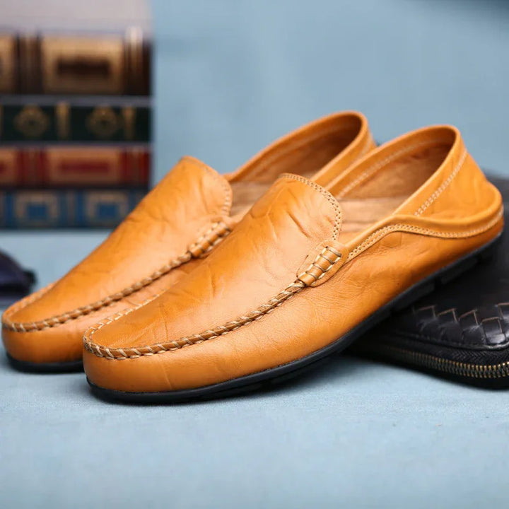 Pair of tan leather men's loafers with braided stitching on a light blue surface and books in the background