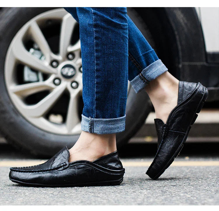 Person wearing black leather loafers and rolled-up blue jeans standing on pavement near car tire