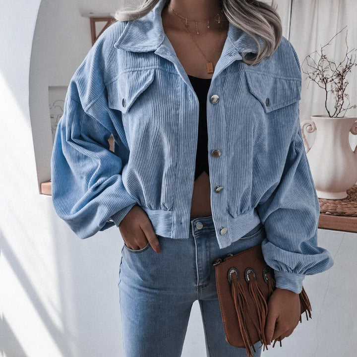 Woman wearing light blue corduroy cropped jacket and blue jeans, holding brown fringe clutch indoors