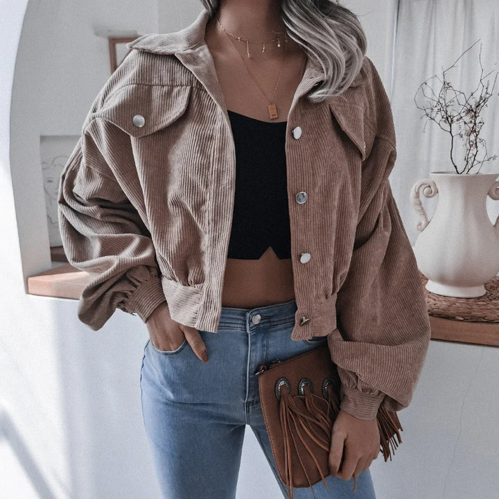Woman wearing cropped beige corduroy jacket with puff sleeves, black crop top, light blue jeans, and holding brown fringed clutch