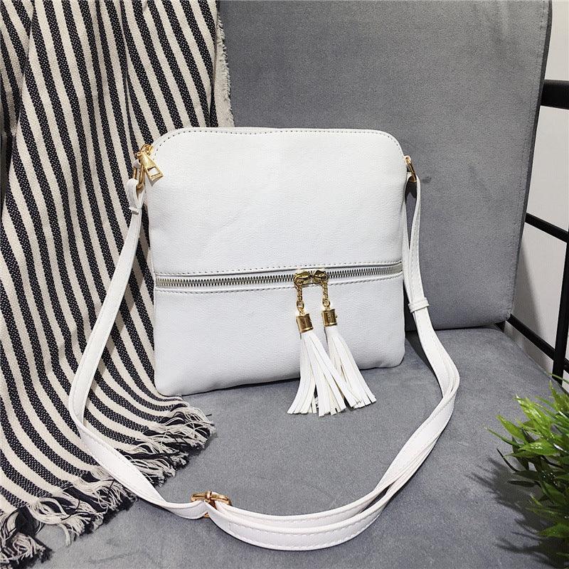 White leather crossbody bag with gold zipper and tassel details on grey chair with striped blanket