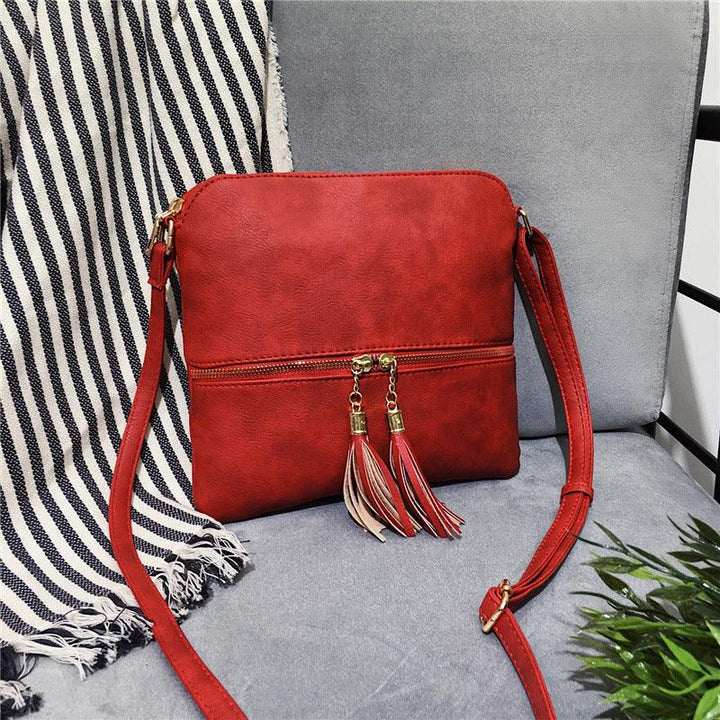 Red leather crossbody bag with tassel zipper pulls on gray chair with striped throw and green plant