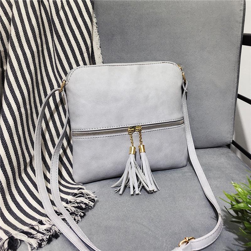 Light gray crossbody bag with gold zipper and tassel detail on gray chair with striped throw