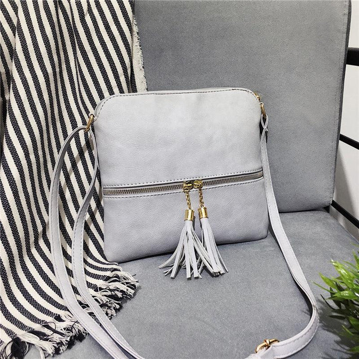 Light gray crossbody bag with gold zipper and tassel detail on gray chair with striped throw