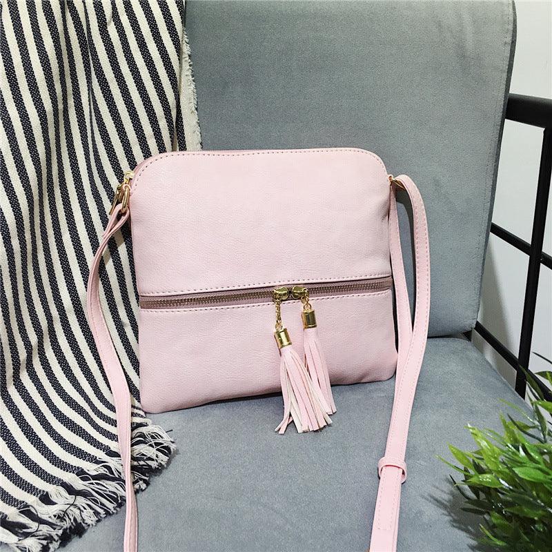 Light pink crossbody bag with gold tassel zipper detail on a gray chair with striped blanket
