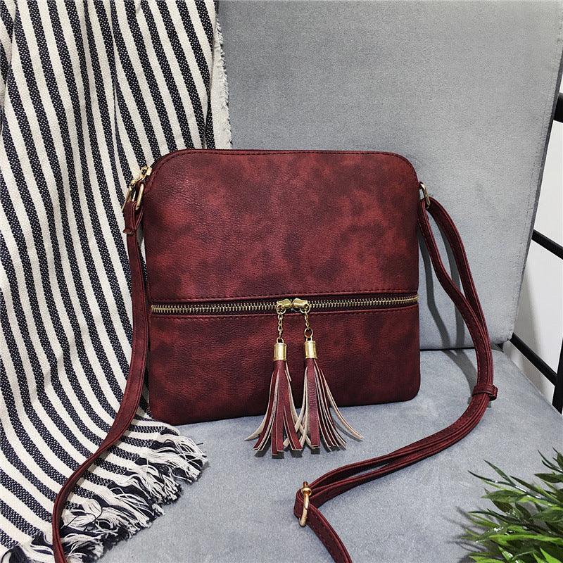 Maroon leather crossbody bag with tassel zipper pulls on gray chair beside striped fabric