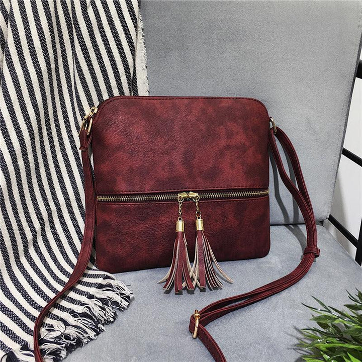 Maroon leather crossbody bag with tassel zipper pulls on gray chair beside striped fabric