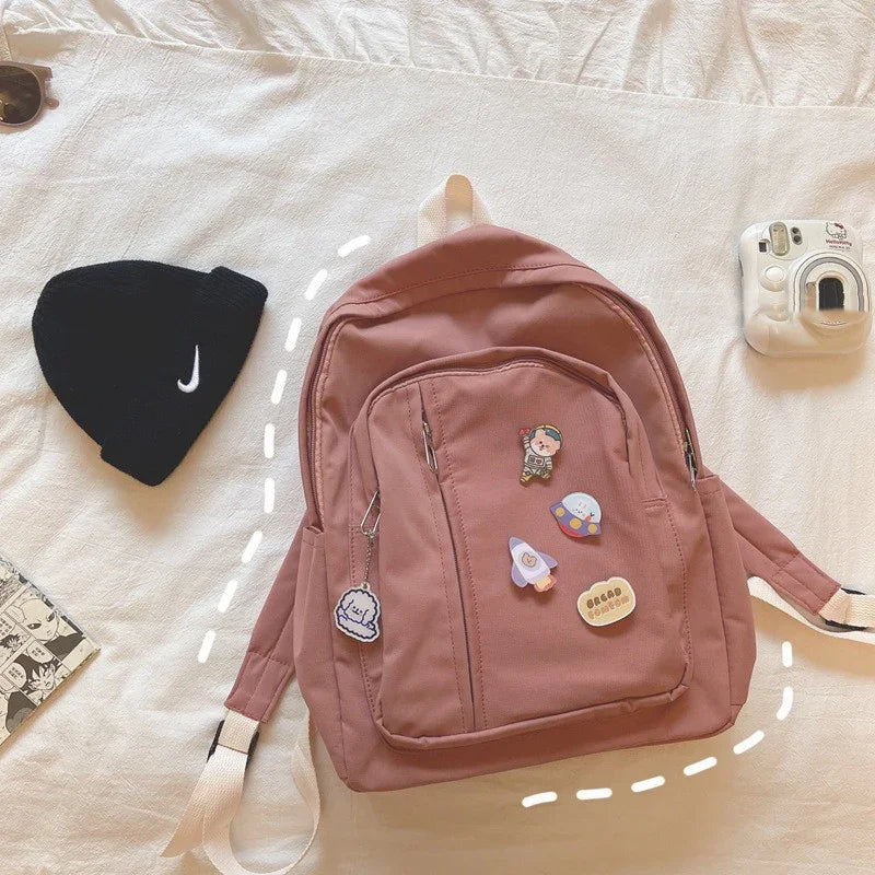 Pink backpack with cute enamel pins, black Nike beanie, and white instant camera on bed