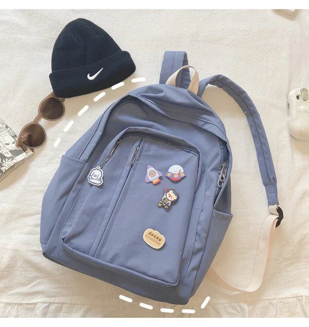 Light blue backpack with cute pins, black beanie, and sunglasses on a white bed