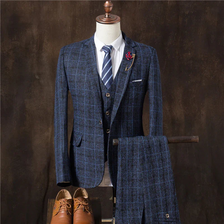 Men's blue checkered three-piece suit with striped tie, white shirt, red lapel pin, and brown leather shoes