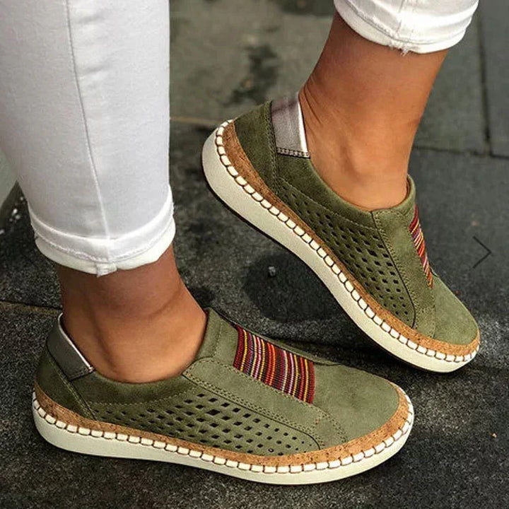 Olive green casual slip-on shoes with perforated sides and multicolor striped elastic detail, worn with white pants