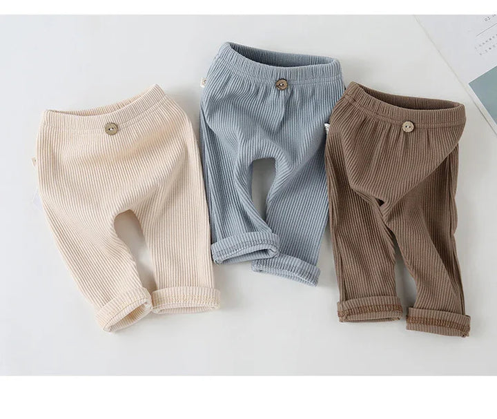 Three pairs of ribbed baby pants in beige, blue, and brown with button details on a white background