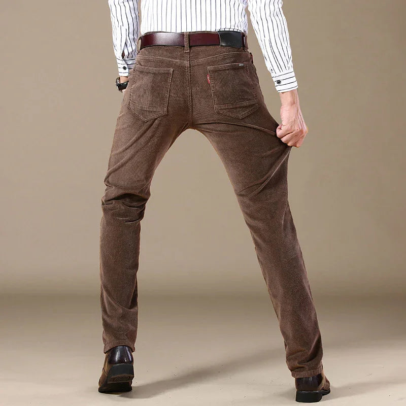 Man wearing brown corduroy pants with a belt, stretching fabric while standing against beige background