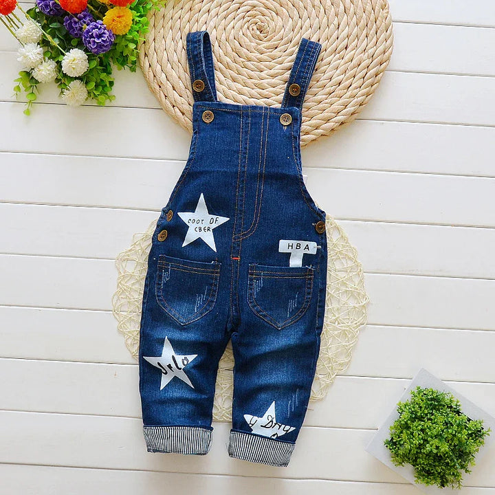 Dark blue denim baby overalls with white star patches and button straps on white wooden floor