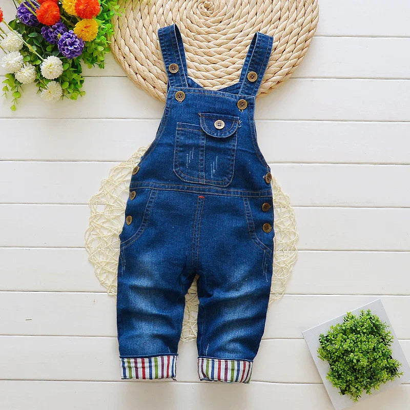 Blue denim toddler overalls with striped cuffs on white wooden floor with flowers and green plant