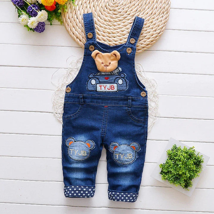 Baby denim overalls with teddy bear patches and TYJB embroidery on front and knees, laid on white wooden floor