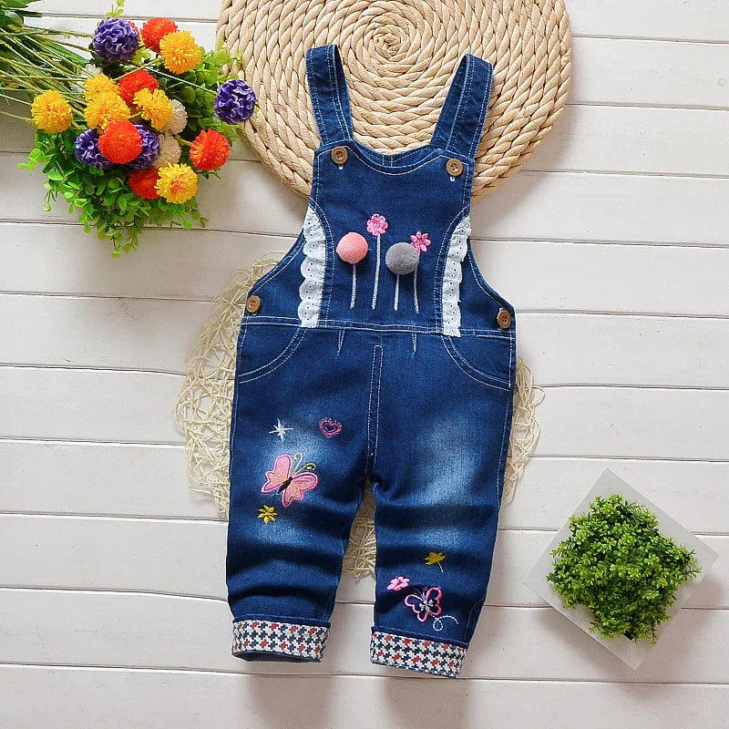 Blue denim baby overalls with embroidered butterflies, flowers, pom-poms, and rolled-up patterned cuffs