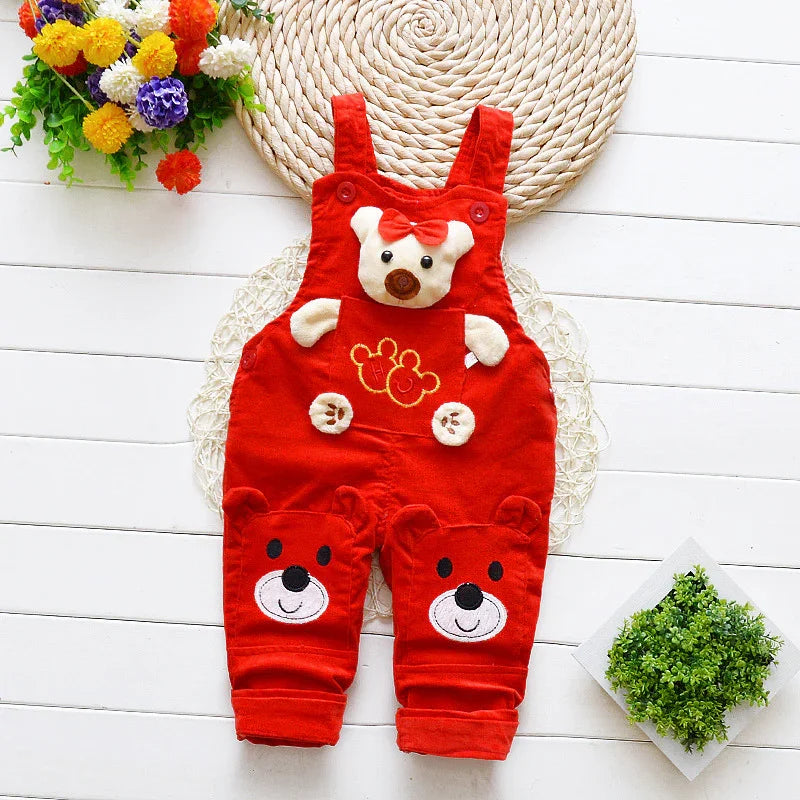Red baby overalls with teddy bear faces and bow detail on wooden floor with flowers and green plant