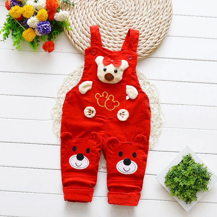 Red baby overalls with teddy bear faces and bow detail on wooden floor with flowers and green plant