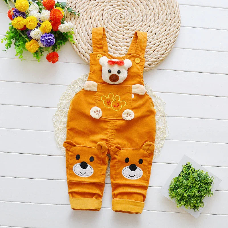 Orange baby overalls with bear faces and paws design, laid flat on white wooden floor with flowers and green plant