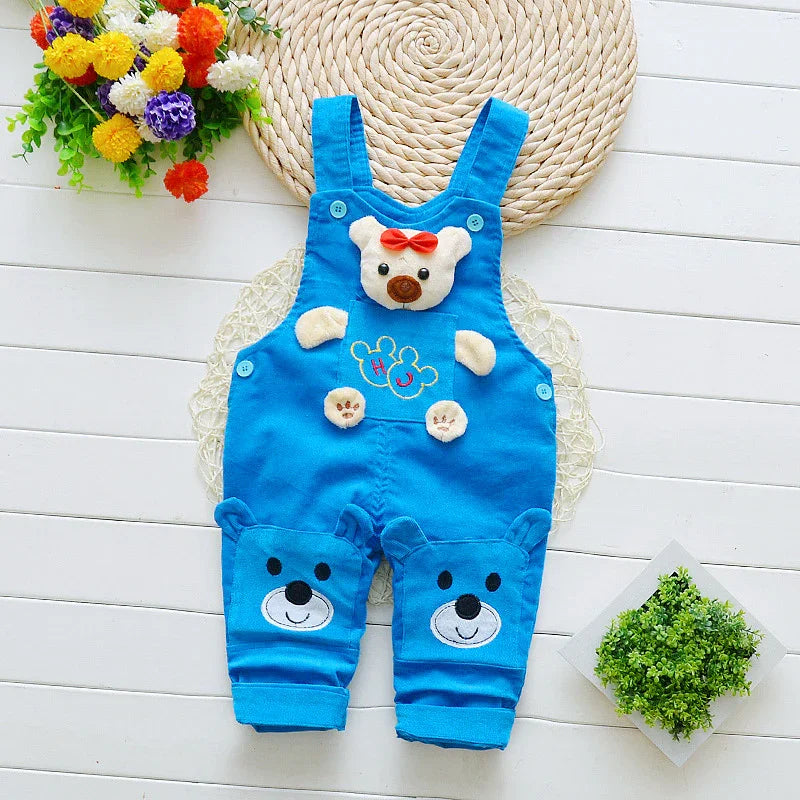 Blue baby romper with teddy bear face and paws design, placed on white wooden floor with flowers and green plant