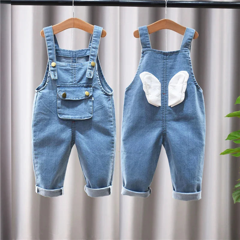 Front and back view of blue denim baby overalls with pockets and white wing patches, hanging on wooden hangers