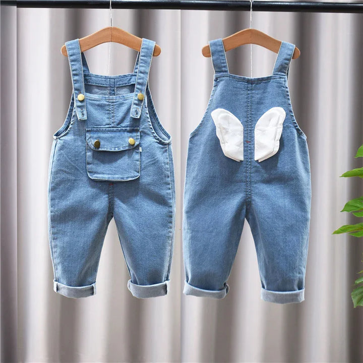 Front and back view of blue denim baby overalls with pockets and white wing patches, hanging on wooden hangers