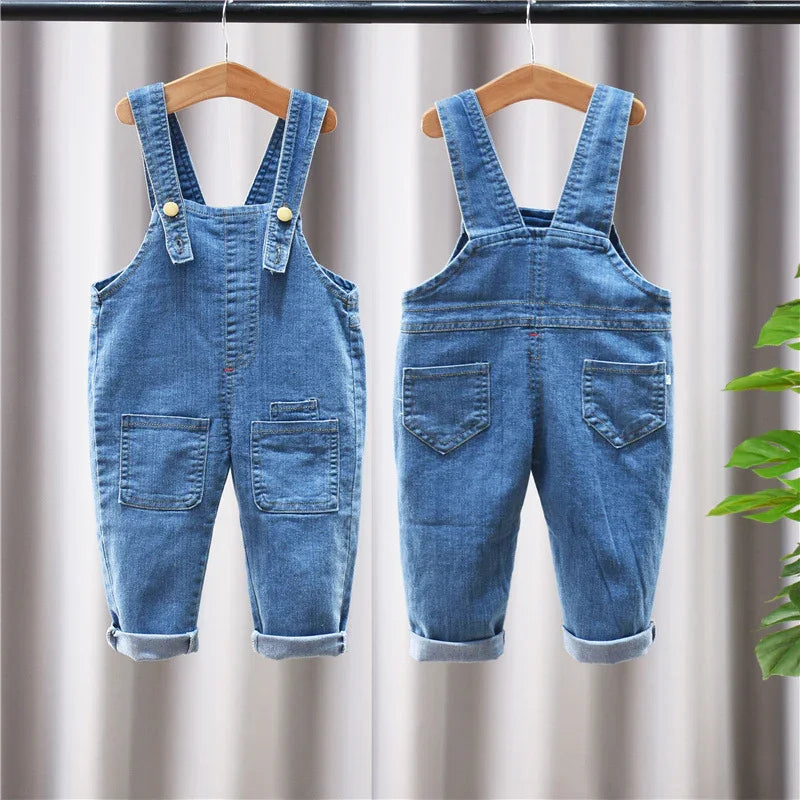 Front and back views of blue denim toddler overalls with rolled cuffs hanging on wooden hangers