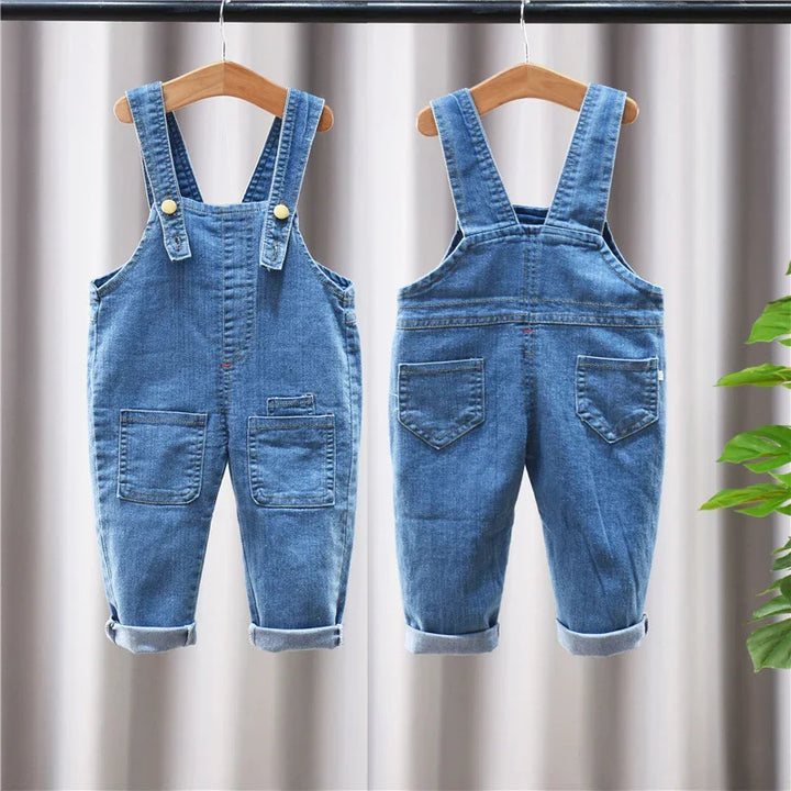 Front and back views of blue denim toddler overalls with rolled cuffs hanging on wooden hangers