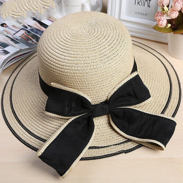 Beige woven straw sun hat with wide black ribbon bow on wooden table with flowers and magazine