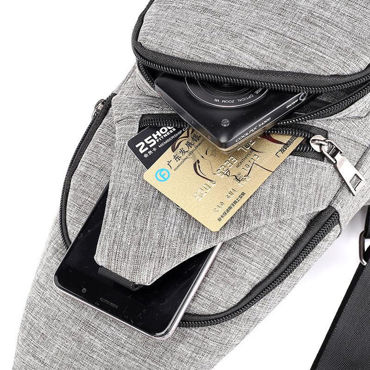 gray fabric sling bag with front zipper pockets holding a black camera, smartphone, and credit cards
