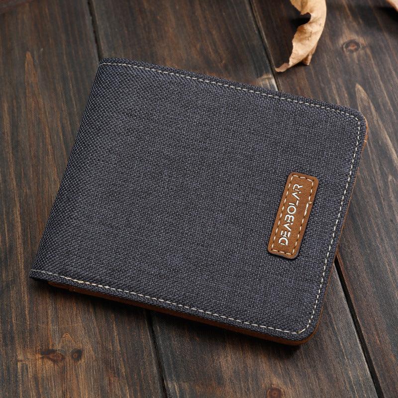 dark gray canvas wallet with brown leather logo patch on wood surface with dried leaves