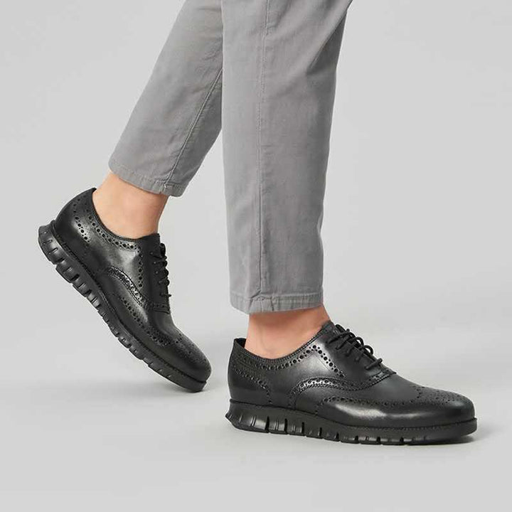 Black leather wingtip brogue shoes with perforated design worn with light gray pants on light gray background