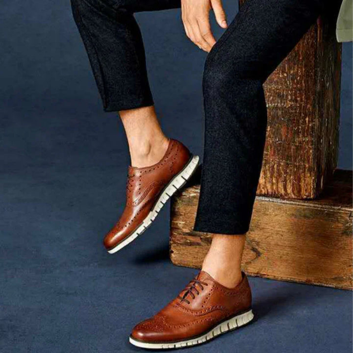 Men's brown leather brogue shoes with white soles worn with black pants on dark floor