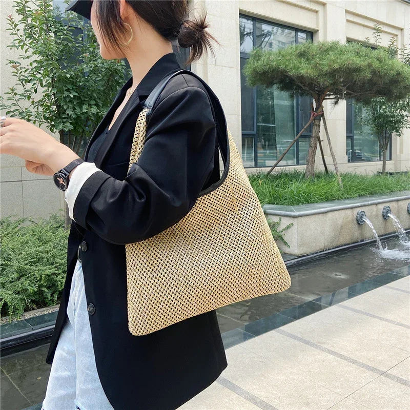 Woman in black blazer carrying a beige woven shoulder bag near modern building and water feature