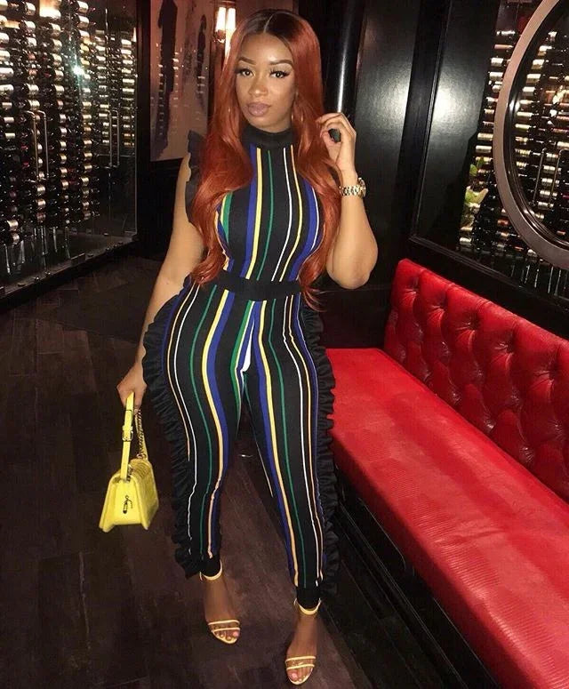 Woman wearing a colorful striped jumpsuit with ruffles, holding a yellow handbag, posing in a stylish lounge with red seating and wine racks