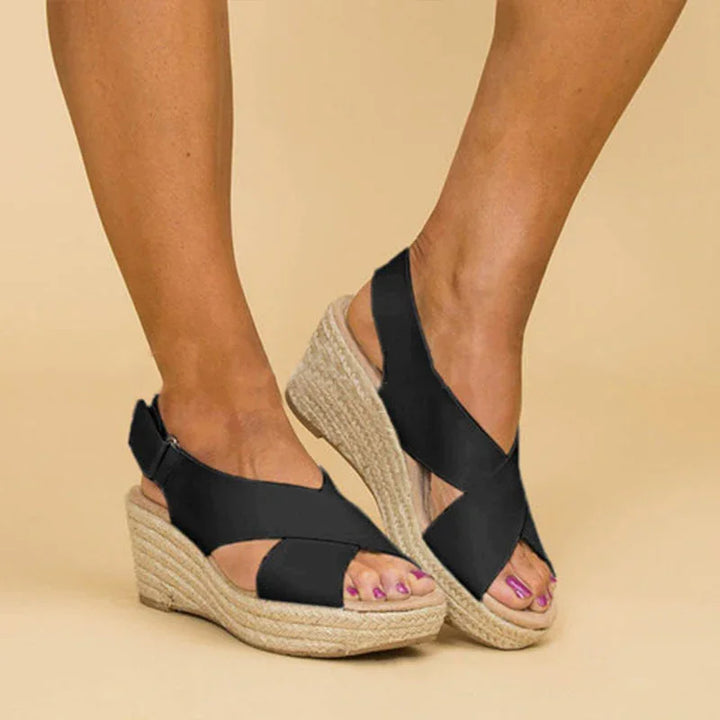 Black wedge espadrille sandals with crisscross straps on feet against beige background